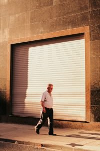 Un homme âgé passe devant un volet roulant fermé par une journée ensoleillée, projetant des ombres.